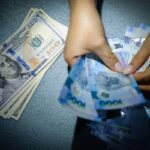 A close-up shot of hands counting Philippine Peso banknotes next to a stack of United States one hundred dollar bills.