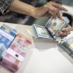 A person counting stacks of US dollars next to bundles of Rupiah banknotes during a currency exchange.