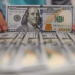 Hands holding a 100 dollar bill over stacks of cash, illustrating the exchange rate between the USD and the Rupiah.