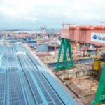 A high-angle view of a vast shipyard facility featuring a large green and orange gantry crane with the Seatrium logo.