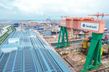 A high-angle view of a vast shipyard facility featuring a large green and orange gantry crane with the Seatrium logo.