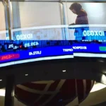 A person walking past a digital stock ticker displaying financial data and market trends related to the JCI at the IDX.