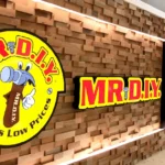 A storefront featuring the yellow and red Mr. DIY logo on a textured wood wall with bright store lighting.